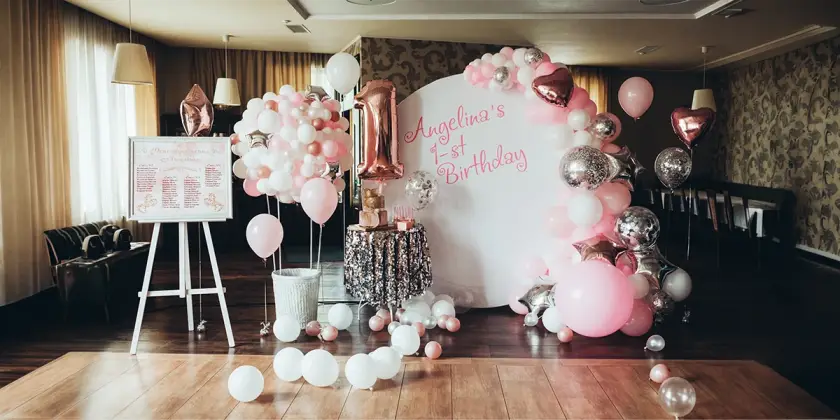 Professional event management setup for a 1st birthday party featuring a white backdrop with pink lettering, a large rose gold number one balloon, an elegant balloon arch, and a sequin cake table.