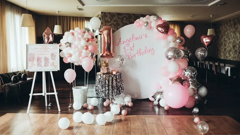Professional event management setup for a 1st birthday party featuring a white backdrop with pink lettering, a large rose gold number one balloon, an elegant balloon arch, and a sequin cake table.