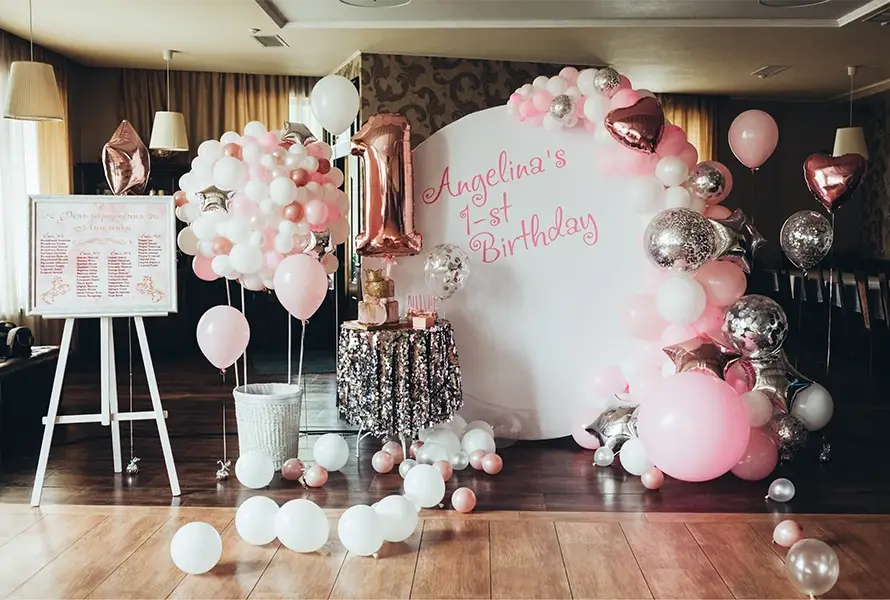 Professional event management setup for a 1st birthday party featuring a white backdrop with pink lettering, a large rose gold number one balloon, an elegant balloon arch, and a sequin cake table.